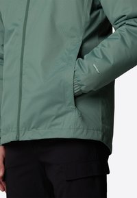 Lys grøn vandtæt jakke med frontlynlås, elastiske manchetter, sidelommer og en struktureret finish. Sorte bukser er delvist synlige.