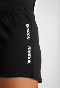 Pantalones cortos deportivos negros con cintura elástica, con logotipos blancos de "Reebok" en los laterales, textura suave y diseño ajustado.