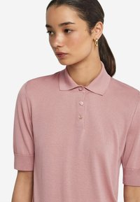 Ljusrosa polo skjorta med korta ärmar, tre knappar på kragen, mjuk textur och en figursydd design. Inga framträdande mönster eller detaljer.