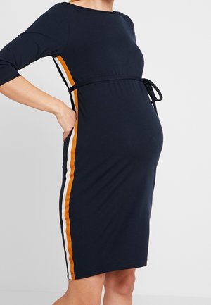 Femme enceinte portant une robe bleu marine ajustée avec des bandes latérales orange et blanches, main reposant sur le bas du dos.