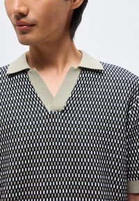 Άνδρας που φοράει μαύρο και λευκό πλεκτό πουκάμισο με μπεζ ανοιχτό γιακά και κοντά μανίκια μπροστά σε μονόχρωμο φόντο.