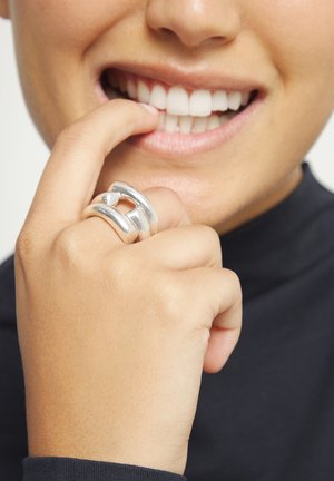 Primer plano de una boca sonriente con dientes blancos y una mano con un anillo grueso de plata tocando suavemente el labio inferior.