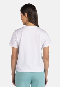 Camiseta de algodón blanca con cuello redondo, mangas cortas y un ajuste relajado. La parte trasera es lisa, sin patrones ni adornos.