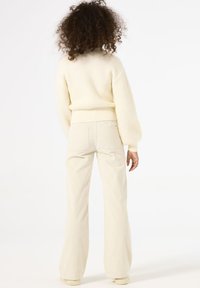Suéter de canalé crema con hombros caídos combinado con pantalones acampanados de un beige claro, que presentan un corte recto y sutiles bolsillos traseros.