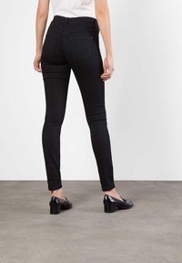 Černé úzké džíny s hladkou texturou, vysokým pasem a dvěma zadními kapsami. Stylizované s černými podpatky na šedém podkladu.