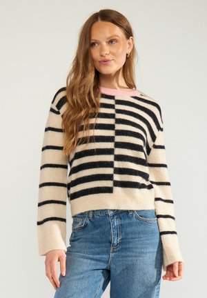Femme aux longs cheveux portant un pull rayé noir et beige avec un col rose et un jean bleu, debout devant un fond clair.