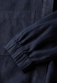 Navyblauwe linnen stof met een close-up van een gathered manchet en een opgestikte zak. De textuur is iets gekreukt met een zachte draping.