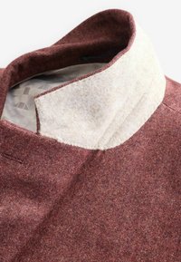 Tissu rouge texturé avec un col beige contrastant. Le col présente des coutures visibles. L'intérieur est doté d'une doublure à motifs.