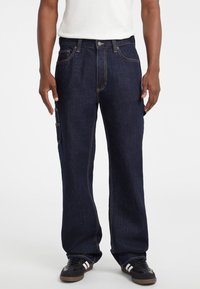 Pantalones cargo de denim azul oscuro con un corte holgado, que cuentan con un cierre frontal de botón, múltiples bolsillos y costuras amarillas en contraste. Zapatillas negras visibles.