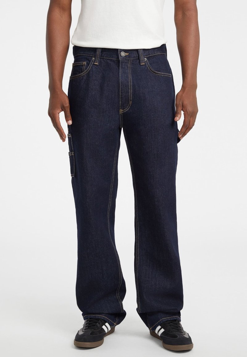 Pantalones cargo de denim azul oscuro con un corte holgado, que cuentan con un cierre frontal de botón, múltiples bolsillos y costuras amarillas en contraste. Zapatillas negras visibles.