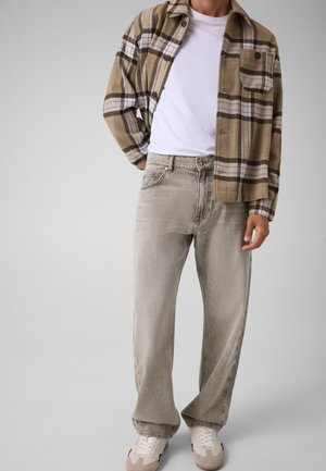 Man draagt een beige geruit overhemd over een wit t-shirt met losse lichtgrijze spijkerbroek en lichtbeige sneakers tegen een effen achtergrond.