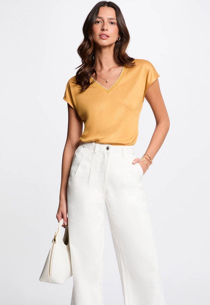 Haut jaune moutarde à col en V avec des manches courtes, associé à un pantalon blanc à jambes larges. Les accessoires comprennent un sac à main blanc. Texture lisse.