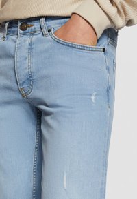 Šviesiai mėlynos spalvos džinsai iš džinsinio audinio su vidutinio aukščio juosmeniu, pažeistais elementais ir kontrastingomis siūlėmis. Klasikinis penkių kišenių dizainas su metaliniais apkaustais.