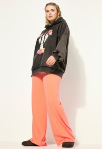 Černá mikina s květinovou grafikou na přední straně; volný střih. Světlé růžové kalhoty s širokými nohavicemi. Model má na sobě tmavé pantofle; celkově hladká textura.