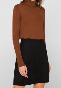 Brauner Rollkragenpullover aus feinem Strickmaterial, kombiniert mit einem schwarzen Wildleder-Minikleid. Das Ensemble hat lange Ärmel und eine figurbetonte Silhouette.