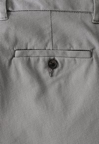 Tissu gris texturé avec une petite poche à rabat et un détail de bouton noir. Le matériau semble tissé et structuré.