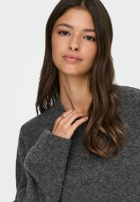 Tumši pelēkas krāsas adīta džemperis ar augstu apkakli un ribotām piedurknēm, kas izceļas ar maigu tekstūru. Roka atpūšas netālu no kakla, demonstrējot vienkāršu gredzenu.