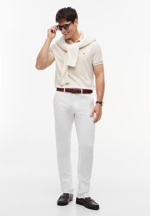 Uomo che indossa una polo beige con maglione sulle spalle, pantaloni bianchi, cintura marrone, mocassini neri, orologio e occhiali da sole, sorride mentre si sistema gli occhiali.