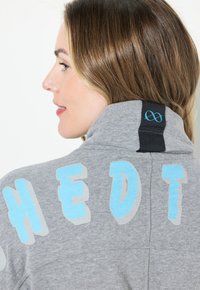 Felpa grigia con colletto alto, caratterizzata da una scritta blu audace e un logo discreto sul colletto. Tessuto morbido e texturizzato con un design aderente.