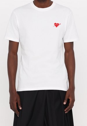Biała bawełniana koszulka z małym graficznym motywem czerwonego serca z strzałą po lewej stronie klatki piersiowej. Krótkie rękawy i okrągły dekolt.