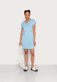 Vestido curto azul claro com decote em V e detalhes de rendas na gola, amarrado na cintura. Acompanhado de uma bolsa branca e sandálias.