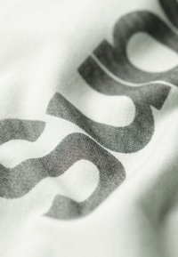 Tela blanca con grandes y audaces patrones de texto en negro. El material parece suave y liso con un ligero brillo. Énfasis en el diseño centrado.