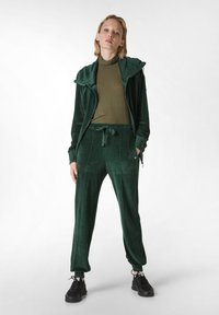 Samt-Trainingsanzug in tiefgrün mit einer Reißverschlussjacke und Kapuze, kombiniert mit passenden Jogginghosen. Schwarze Sneaker runden den Look ab.