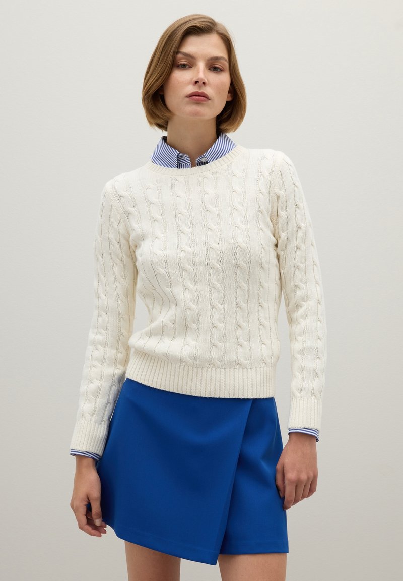 Cremfarbener Zopfmuster-Pullover mit geripptem Saum, über einem kragenbehafteten blau-weiß gestreiften Hemd getragen, kombiniert mit einem blauen Rock.