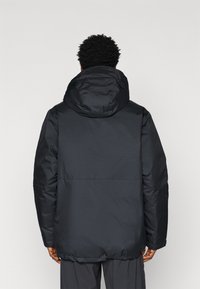 Jachetă pufă neagră cu glugă detașabilă, având o textură netedă și o croială relaxată. Include o cusătură contrastantă la nivelul taliei.