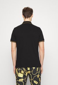 Svart polo skorta med gult kantband på kragen och ärmarna. Buren med mönstrade svarta byxor som har guldkedje-mönster.