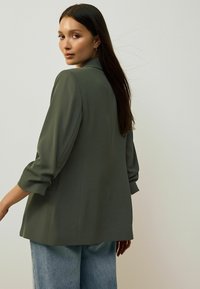 Olivově zelený blazer se strukturovanými rameny, rukávy po lokty a volným střihem, kombinovaný s světle modrými širokými džíny. Hladká textura látky.