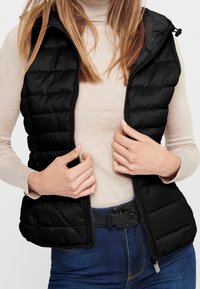 Chaleco de pluma negro con textura acolchada, cremallera frontal y capucha. Lleva puesto un suéter de cuello alto beige claro, combinado con unos vaqueros y un cinturón negro.