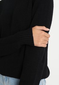 Zwarte trui met ribgebreide boorden, zachte textuur en een relaxte pasvorm, gedeeltelijk getoond met een hand die over de arm kruist.