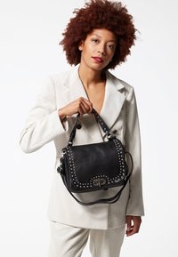 Borsa a tracolla in pelle nera con dettagli borchiati e chiusura con logo. Presenta una tracolla lunga e una superficie testurizzata, indossata da una modella in un completo beige.