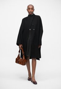 Zwarte wollen mantel met hoge kraag en dubbele knoopsluiting, gecombineerd met een donkere rok en een bruine leren handtas. Paarse ballet flats maken de look af.