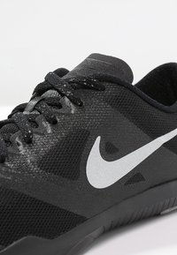Scarpa sportiva nera con una tomaia in rete testurizzata, stringhe rotonde e un grande logo Nike swoosh argento sul lato.