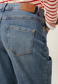 Jeans de denim azul con un parche de cuero en la cintura, bolsillo trasero cosido y un ajuste ligeramente holgado con un acabado texturizado.