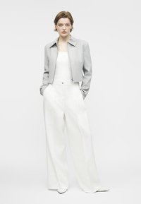 Frau mit kurzen Haaren, die eine hellgraue, kurz geschnittene Jacke, ein weißes Oberteil, weite weiße Hosen und weiße spitze Schuhe trägt, steht mit Händen in den Taschen.