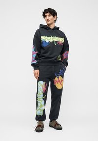 Mann trägt schwarzen Hoodie und Jogginghose mit bunten Graffiti-Designs, steht mit Händen in den Taschen und schaut zur Seite.