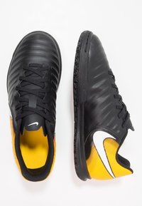 Zapatillas de fútbol sintéticas negras con superficie texturizada, detalles amarillos y logotipo de Nike en blanco. Cuenta con un diseño con cordones y suela de goma.