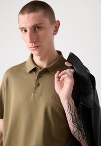 Polo shirt vert olive avec deux boutons et un logo CK, tenu par un mannequin portant une veste en cuir noire sur une épaule ; tatouage sur l'avant-bras.