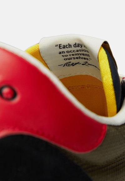 Zapatilla de cordones con punta roja, cuerpo de tela gris, forro amarillo y una cita impresa en la lengüeta interior.