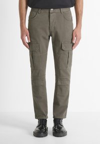 Uomo che indossa pantaloni cargo verde oliva con tasche a patta su cosce e ginocchia, abbinati a stivali neri in pelle con lacci, in piedi contro uno sfondo bianco.