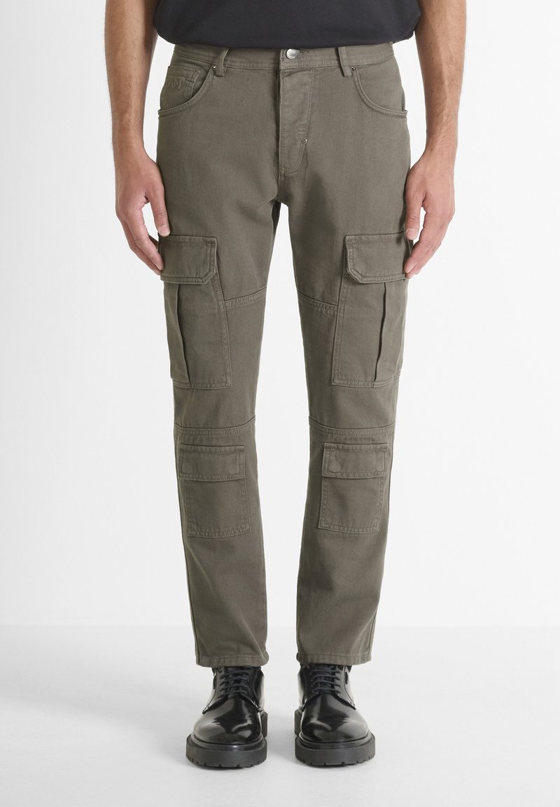Uomo che indossa pantaloni cargo verde oliva con tasche a patta su cosce e ginocchia, abbinati a stivali neri in pelle con lacci, in piedi contro uno sfondo bianco.