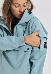 Giacca a vento azzurro chiaro con chiusura a zip, cordini regolabili e tasca a toppa sulla manica. Tessuto liscio e polsini.