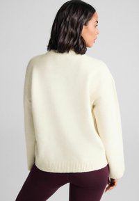 Kræmfarvet sweater lavet af blødt stof, med en afslappet pasform, ribbede kanter og dropped skuldre. Bæres med tætsiddende mørke leggings.