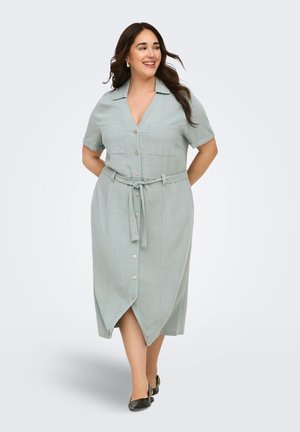 Femme marchant dans une robe midi vert clair avec boutons sur le devant, manches courtes, col, poches avant, ceinture et chaussures plates noires.