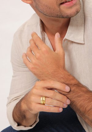 Gold-toned ring with a textured, hammered design worn on a hand. Light-colored linen shirt and visible skin texture on the arm.