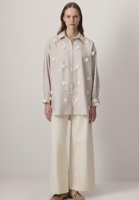 Camisa beige clara con apliques florales, diseño de botones y mangas largas, combinada con pantalones de pierna ancha color crema y calzado claro.