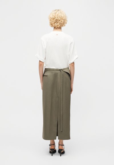 Άτομο με ξανθά σγουρά μαλλιά που φοράει λευκή κοντομάνικη μπλούζα και μακριά ελιάς πράσινη φούστα με ζώνη, ορατό από πίσω.